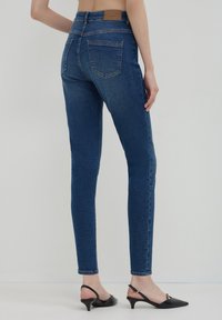 Femme portant un jean skinny bleu foncé et des escarpins à talons bas noirs, vue de dos sur un fond clair uni.