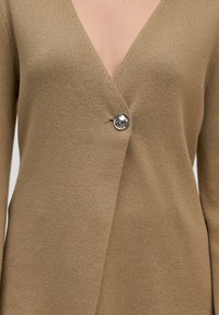 Beige gebreide cardigan met een diepe V-hals, voorzien van een zilveren knoopaccent aan de linkerkant en een gladde, zachte textuur.