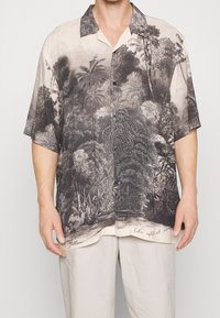 Uomo in camicia a maniche corte con bottoni, decorata con un dettagliato motivo di foresta tropicale in bianco e nero, e pantaloni beige, in piedi di fronte a uno sfondo semplice.