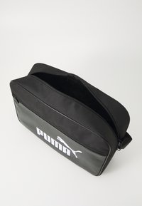 Borsa nera Puma con texture liscia, grande logo bianco, chiusura con zip e tracolla regolabile. Design rettangolare, adatta per l'uso quotidiano.