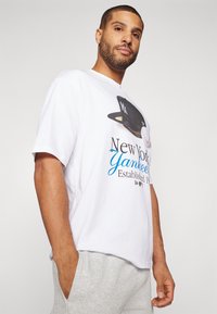 Camiseta blanca de algodón con un gráfico de una gorra negra de los New York Yankees, texto en azul y negro, y un ajuste casual con mangas cortas.