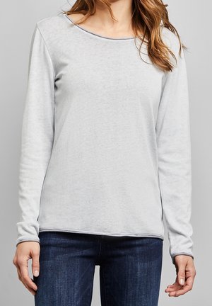 Femme portant un haut à manches longues gris clair et un jean bleu foncé, debout devant un fond gris uni.