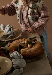 Ciasto Bundt o złocisto-brązowej skórce, udekorowane owocami, serwowane na drewnianej desce z szarymi lnianymi serwetkami i ceramicznymi talerzami w pobliżu.