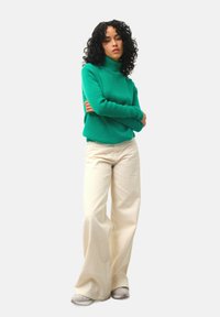 Maglione verde a collo alto in un morbido filato, abbinato a pantaloni a gamba larga color crema e sneakers chiare, con il modello in piedi con le braccia incrociate.