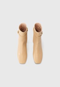 Beige leren enkellaarsjes met een gladde afwerking, vierkante neus en decoratieve goudkleurige hardware bij de enkel. Geen patronen of versieringen.