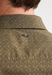 Cuello de camisa color verde oliva con un patrón geométrico texturizado, que presenta una pequeña etiqueta de logo plateada unida cerca del cuello.