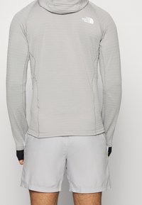 Camiseta de manga larga gris clara con capucha, textura suave, costuras planas y un pequeño logo blanco. Combinada con shorts gris claros. Diseño de ropa activa casual.