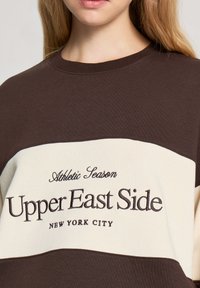 Felpa marrone e crema con il testo "Athletic Season" e "Upper East Side" ricamato in marrone scuro, con colletto a costine.