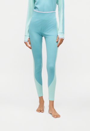 Femme portant un legging bleu bicolore taille haute et un haut à manches longues assorti, debout pieds nus sur un fond blanc.