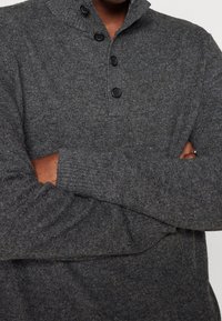 Person mit verschränkten Armen, die einen dunkelgrauen, knöpfbaren Strickpullover mit Stehkragen trägt, wobei die strukturierten Stoffdetails sichtbar sind.