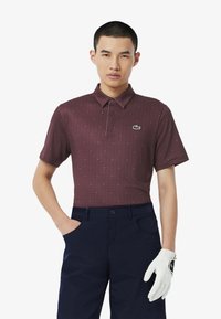 Burgundowa koszulka polo z krótkim rękawem w geometryczny wzór oraz zielonym logo krokodyla, zestawiona z granatowymi spodniami i białą rękawicą golfową.