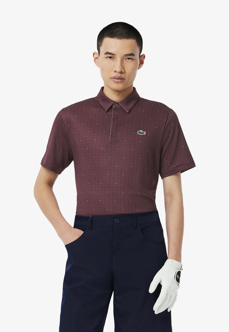 Burgundowa koszulka polo z krótkim rękawem w geometryczny wzór oraz zielonym logo krokodyla, zestawiona z granatowymi spodniami i białą rękawicą golfową.