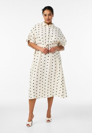 Naine seisab seljas valge poolpikk kleit mustade täppidega, vöödiga talje ja lühikeste varrukatega, jalas valged kontsaga sandaalid.