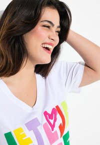 T-shirt blanc à col en V avec des lettres en feutre colorées épelant "LAISSEZ L'AMOUR" dans des couleurs variées, avec un accent en forme de cœur rose.