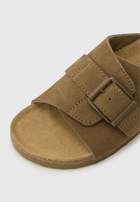 Sandale ouverte en daim marron avec une large bride et une boucle réglable sur un fond gris clair.