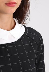 Musta paita, jossa on valkoinen ruudukko-kuvio, pyöreä pääntie ja valkoinen kaulus. Sileä pinta ja rento muotoilu.