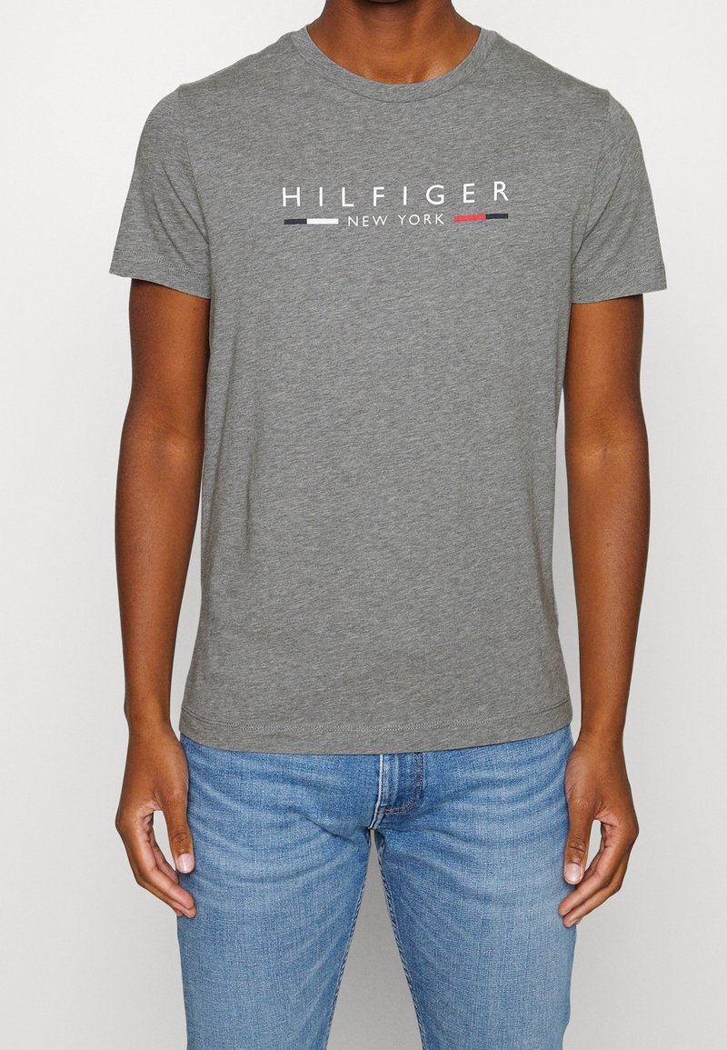Man som bär en grå t-shirt med kort ärm med texten "HILFIGER NEW YORK" och logobars, i kombination med ljusblå jeans.