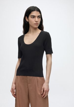 Femeie cu păr lung și închis la culoare, purtând un top negru cu mâneci scurte, cu dungi, și pantaloni largi de culoare maro deschis, stând în picioare pe un fundal simplu.