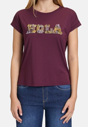 Vrouw draagt een paarse T-shirt met korte mouwen en het woord "DOLA" in getextureerde bloemletters, gecombineerd met een blauwe spijkerbroek.