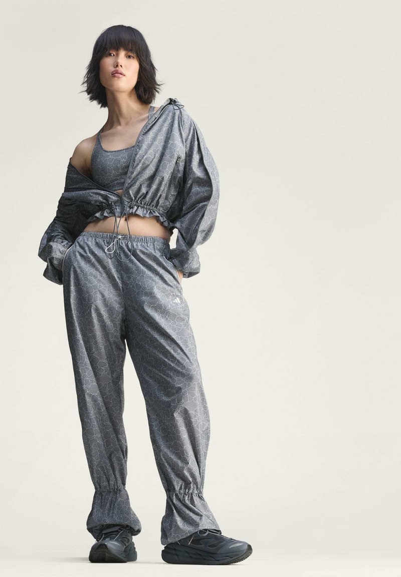 Woman wearing gray patterned sports bra, matching track jacket and pants with black sneakers, posing with hands in pockets against plain background.