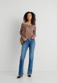 Γυναίκα με σγουρά μαλλιά που φοράει μπλούζα καρό με ακάλυπτους ώμους, μπλε τζιν, μαύρες γόβες και ταμπά τσάντα ώμου, στέκεται μπροστά σε λευκό φόντο.