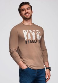 Braunes Langarm-T-Shirt mit einem weißen und schwarzen Grafikdruck, auf dem "NYC" und "BROOKLYN" steht, kombiniert mit blauen Jeans.
