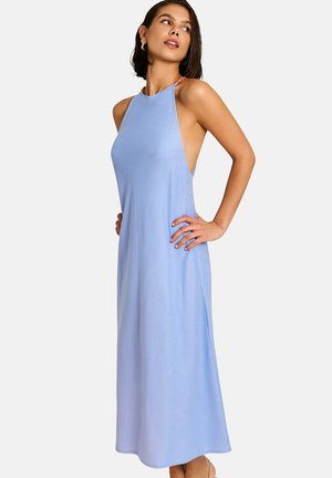 Femme portant une robe longue sans manches bleu clair avec un col licou, debout les mains sur les hanches, regardant sur le côté.