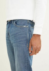 Vaqueros de mezclilla de color azul claro, con corte slim, bolsillos delanteros y una pequeña etiqueta con logo. Combinados con una camisa blanca de manga larga.