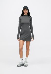 Pitkähihainen harmaa urheilumekko, jossa on korkea kaulus ja sivuilla lävistysyksityiskohtia. Yhdistetty mustaan lippalakkiin ja harmaisiin jalkineisiin.