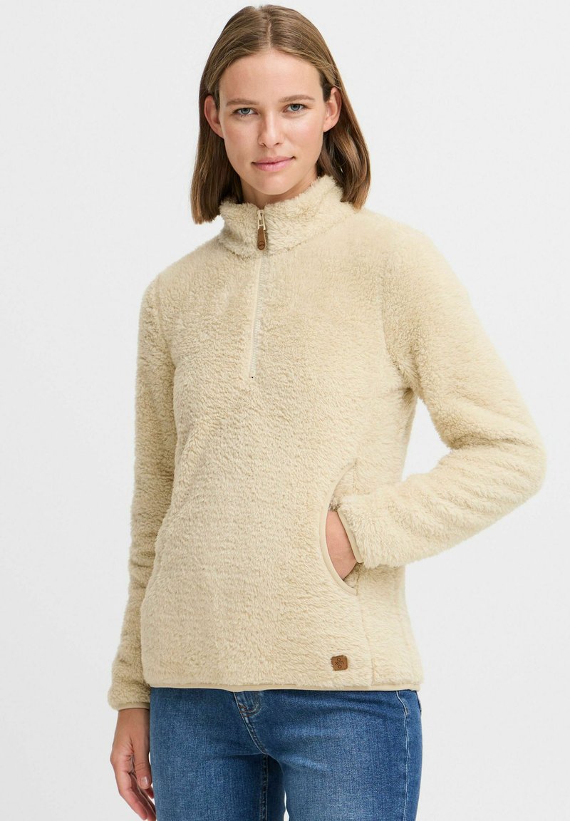 Beige fleece pullover with a high collar and half-zip, featuring side pockets and a textured surface. Small branded badge at the hem.