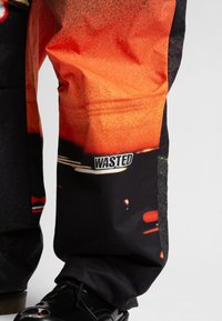 Čierne nohavice s oranžovým grafickým vzorom, s viditeľnou štítkou "WASTED". Materiál sa zdá byť odolný a mierne texturovaný.