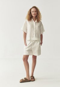 White linen short-sleeve shirt with buttons and matching shorts. Brown sandals with adjustable strap. Soft texture and relaxed fit.