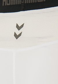 Pantaloni corti sportivi bianchi con una cintura testurizzata nera, caratterizzati da un design con logo e due chevron rivolti verso il basso in grigio.