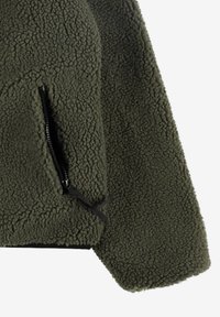 Kurtka polarowa w kolorze oliwkowej zieleni o teksturowanej powierzchni, z czarną kieszenią zapinaną na zamek oraz prostym obszyciem na dole.