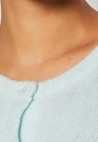 Cardigan azul claro con una textura suave, con cuello redondo y un único cierre de botón blanco en el escote.