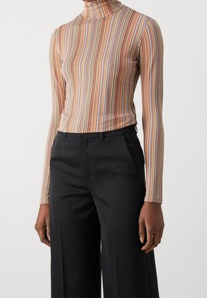 Personne portant un col roulé rayé multicolore à manches longues et un pantalon noir taille haute à jambes larges, se tenant devant un fond uni.