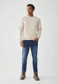 Beige genser med rund hals, myk tekstur, i kombinasjon med blå jeans og brune snørestøvler; stående pose mot en lys bakgrunn.