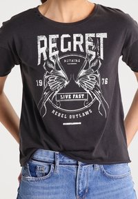 Camiseta de algodón negra con un gráfico impreso en serigrafía que incluye texto y elementos decorativos, como alas y armas de fuego, en blanco.