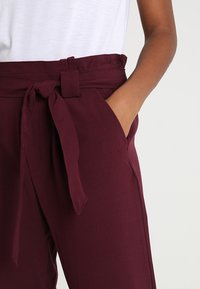 Wijnkleurige broek met een strikband, zijzakken en een gladde stoftextuur. Gecombineerd met een eenvoudig wit bovenstuk.