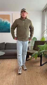 Flis pulover s četvrtastim patentnim zatvaračem u maslinasto zelenoj boji, s prednjim džepom, upareno s svijetloplavim trapericama s manšetama i bež tenisicama na teksturiranom tepihu.