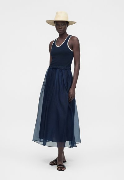 Γυναίκα με μπεζ καπέλο με φαρδύ γείσο, μπλε αμάνικη μπλούζα με λευκή τρέσα, διάφανη μπλε midi φούστα και μαύρα σανδάλια ανοιχτά στα δάχτυλα, στέκεται σε λευκό φόντο.