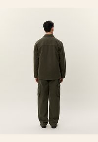 Uomo in piedi con le spalle rivolte, che indossa una giacca verde oliva e pantaloni cargo abbinati, su uno sfondo beige semplice.