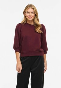 Femme aux longs cheveux blonds portant un haut bordeaux à manches bouffantes et un pantalon noir, debout devant un fond blanc uni.