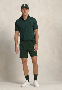Tricou polo verde închis cu guler, asortat cu șorturi corespunzătoare. Purta cu șosete verzi închise și adidași albi. Prezintă un logo pe tricou.