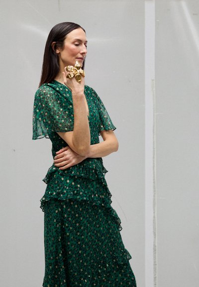 Vrouw in groene jurk met luipaardprint poseert met hand bij gezicht, draagt een oversized gouden bloemenring tegen een effen grijze achtergrond.