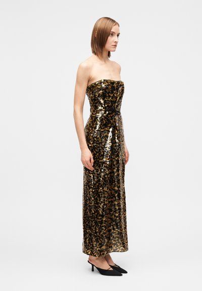 Woman in a strapless, ankle-length sequined dress with black and gold abstract pattern, wearing black pointed-toe heels, standing sideways.