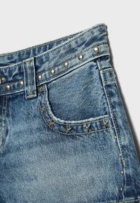 Gros plan sur une poche de jean en denim bleu avec des clous métalliques argentés ronds et en forme d’étoile le long du bord de la poche et de la ceinture.