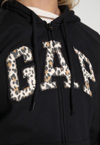 Felpa nera con logo "GAP" in stampa leopardata, con cuciture a contrasto e cordini regolabili sul cappuccio. Materiale in morbido cotone misto.