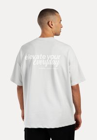 Camiseta de gran tamaño en gris claro con mangas cortas, que presenta texto en blanco que dice "eleva tu día a día" en la parte posterior, con una textura de tela suave.