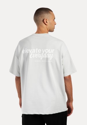 Camiseta de gran tamaño en gris claro con mangas cortas, que presenta texto en blanco que dice "eleva tu día a día" en la parte posterior, con una textura de tela suave.